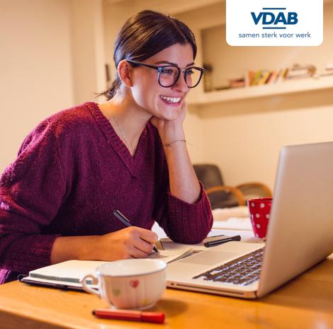 Jonge vrouw met bril en pen in de hand kijkt naar laptop aan haar bureau.